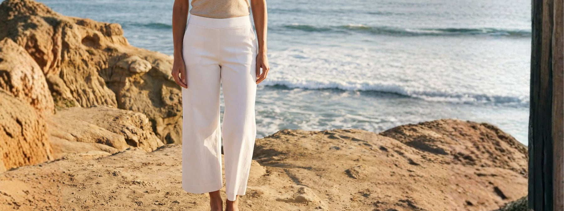 Person wearing a beige top and white pants standing on a rocky beach with ocean waves in the background.