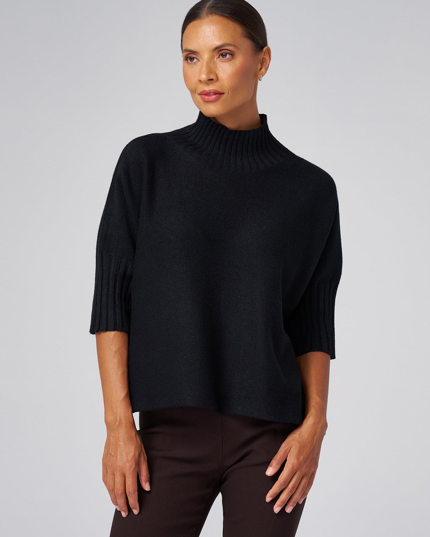 A woman wearing a black ribbed mock-neck knit sweater with three-quarter sleeves, paired with dark brown trousers, against a solid light gray background.
