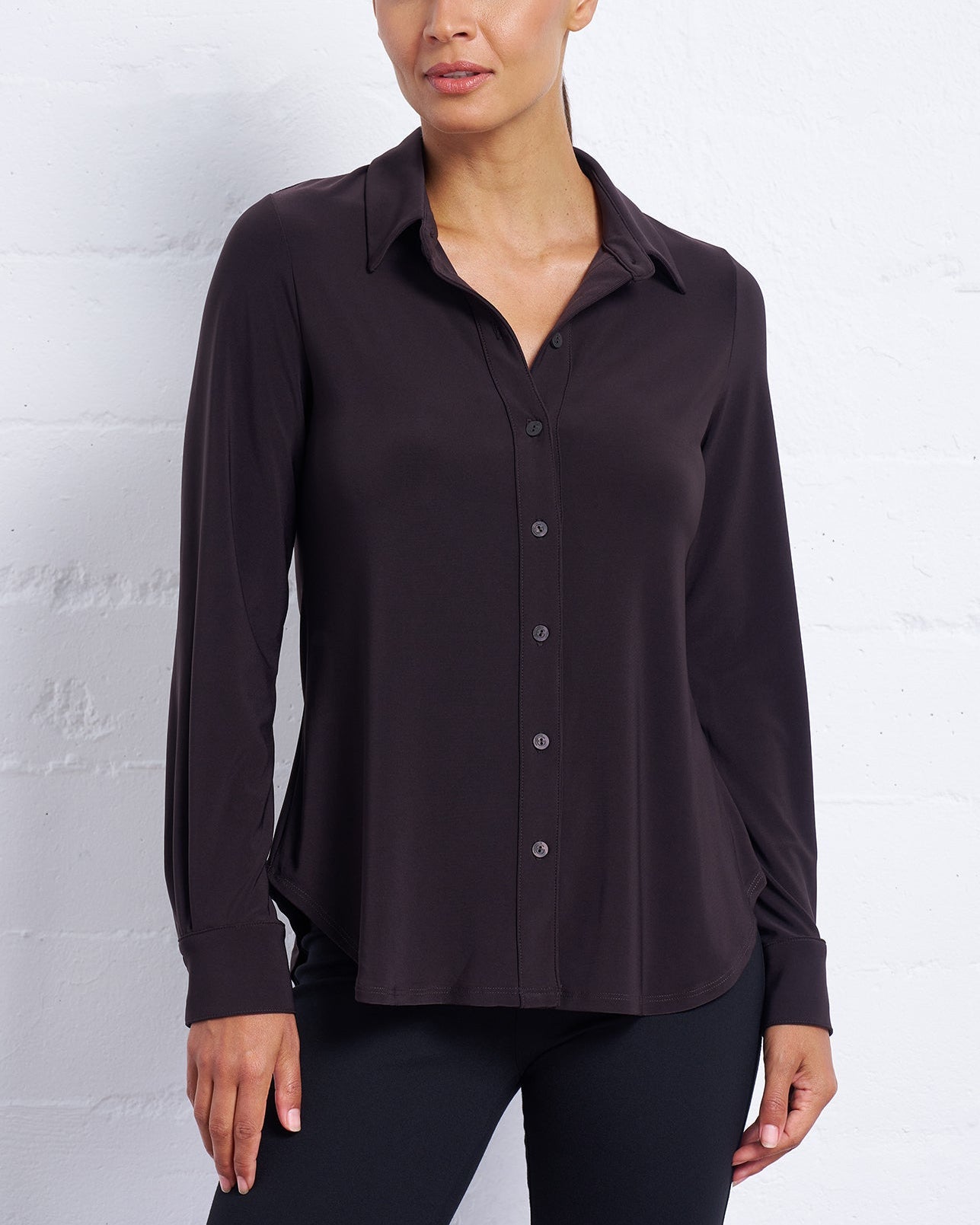Woman wearing a dark brown button-up shirt against a white brick wall.
