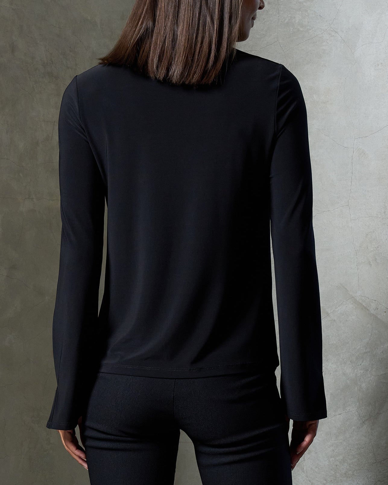 A woman shown from the back wearing a black long-sleeve knit top and matching black pants against a textured gray studio backdrop.
