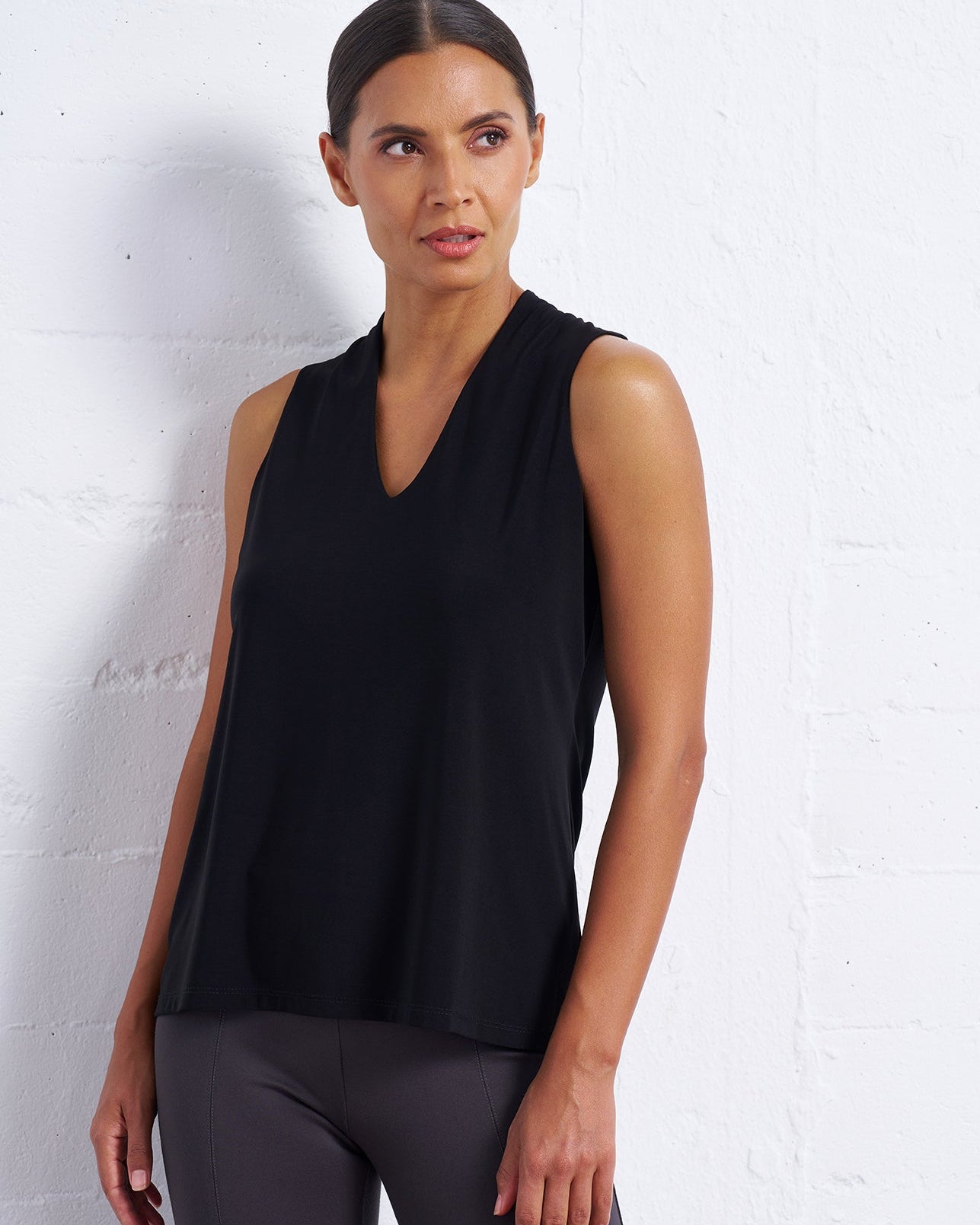 A woman modeling a sleeveless black V-neck top and dark gray leggings against a white brick wall.