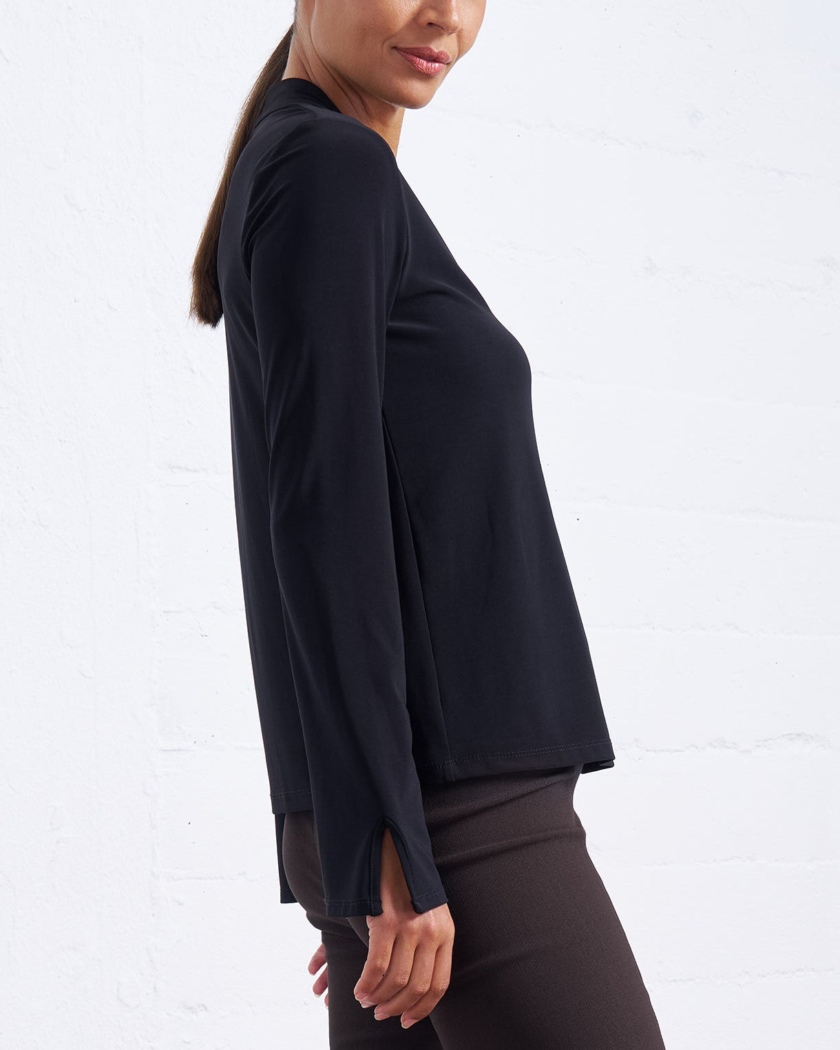 A woman modeling a black long-sleeve crop top with side slits, paired with dark brown pants, standing against a white brick wall.