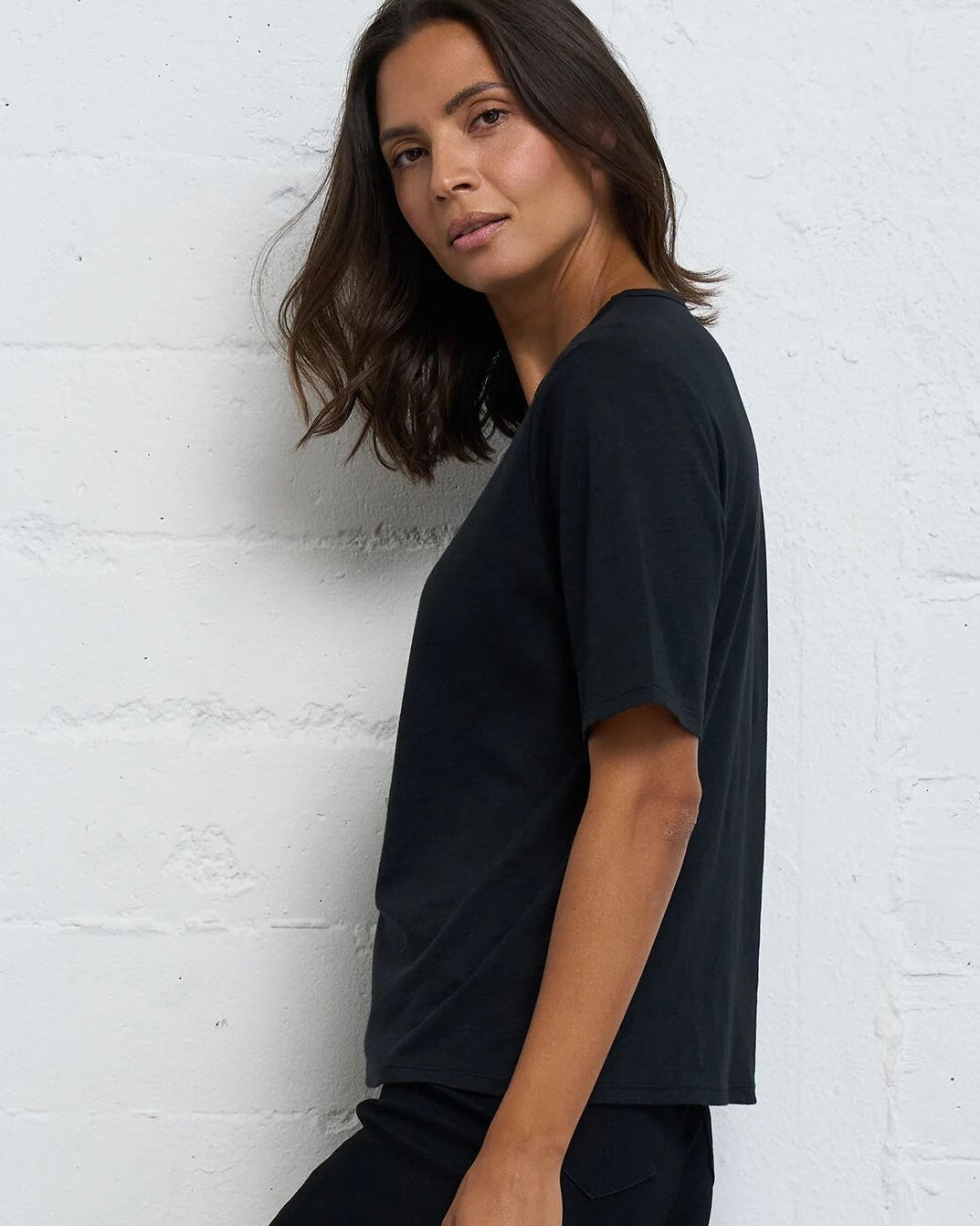 A woman wearing a black short-sleeve crewneck t-shirt and black pants, posing against a white painted brick wall.