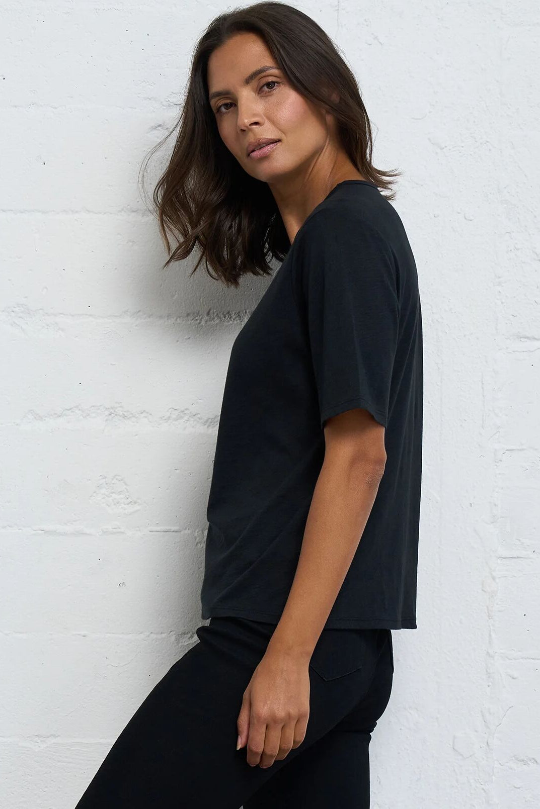 A woman wearing a black short-sleeve crewneck t-shirt and black pants, posing against a white painted brick wall.